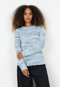 Femeie cu păr creț purtând un pulover tricotat cu dungi albastre și gri și pantaloni negri, pozând cu un braț peste corp.