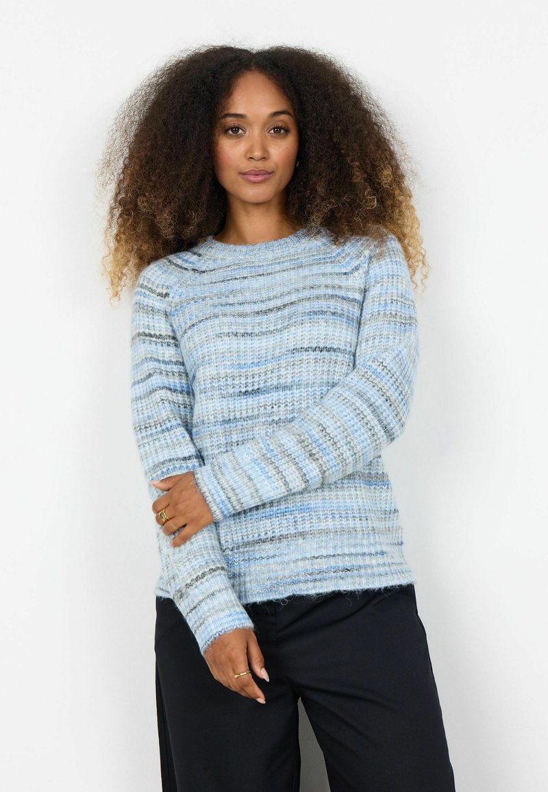 Femeie cu păr creț purtând un pulover tricotat cu dungi albastre și gri și pantaloni negri, pozând cu un braț peste corp.