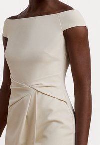 Off-shoulder klänning i krämfärg, tillverkad av mjukt tyg, med en figurnära överdel och draperade detaljer vid midjan, vilket skapar eleganta fickor.