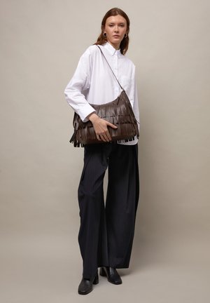 Žena v bílé košili s knoflíky a širokými černými kalhotami držící velkou hnědou kabelku s třásněmi na rameni, na jednobarevném béžovém pozadí.