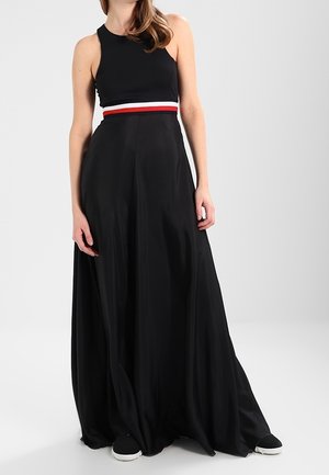 Zwarte maxi-jurk met een nauwsluitend mouwloos lijfje en een contrasterende rood-witte tailleband. Vloeiend materiaal reikt tot de vloer. Witte sneakers zichtbaar.