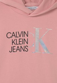 Sudadera rosa con texto negro "Calvin Klein Jeans" y gran logotipo plateado reflectante "K" en la parte frontal, con capucha y etiqueta de tela en el interior.