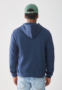 Mann som vender bort, iført en grønn Polo-kappe, marineblå hettegenser med hetta ned, og lyseblå jeans med synlige baklommer.