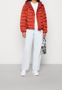 Person som bär en rostorange puffjacka, vit T-shirt, ljusblå jeans, vita sneakers och håller en handväska med svartvit mönster.
