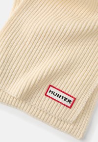Krémová rebrovaná čiapka vyrobená z pleteného materiálu, s obdĺžnikovou nášivkou s nápisom "HUNTER" v čiernej a červenej farbe.