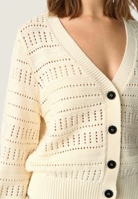 Virkat cardigan i krämvit med djup V-ringning, med vertikala utskärningsmönster och svarta knappar längst fram. Mjuk textur.