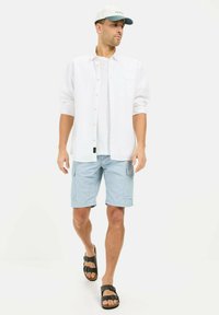 Lichte witte button-up shirt, witte t-shirt, lichtblauwe cargo shorts, zwarte sandalen met twee gespen en een lichtblauwe pet.
