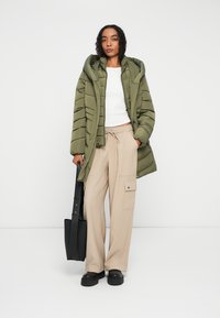 Olivengrønn polstret jakke med hette, hvit crop topp, beige vide cargo bukser og svarte platåskjær, holder en svart skulderveske.