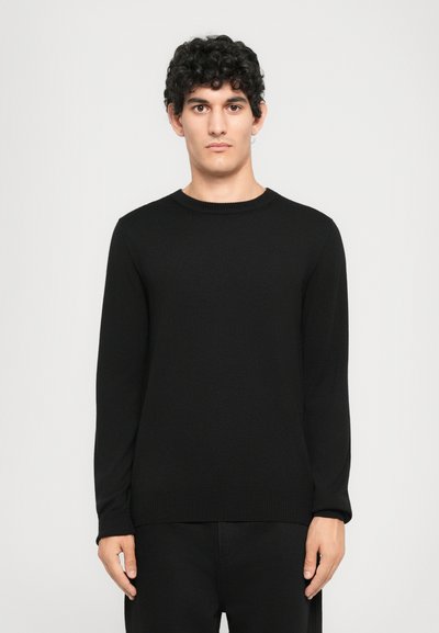 Giovane con capelli ricci che indossa un maglione lungo nero semplice e pantaloni neri su uno sfondo grigio chiaro, rivolto frontalmente.