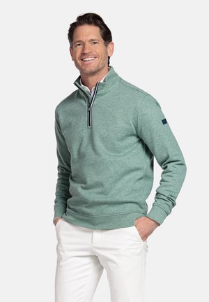 Lachende man met bruin haar, gekleed in een groen sweatshirt met een kwartrits en witte broek, handen in de zakken, staand tegen een effen achtergrond.