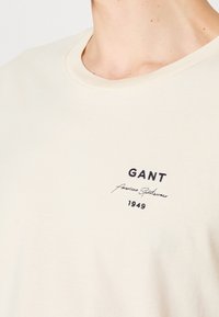 Gros plan sur un t-shirt beige clair à col rond avec l'inscription brodée "GANT American Sportswear 1949" sur la poitrine.