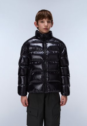 Garçon portant une doudoune noire brillante avec un col montant et un pantalon cargo noir, debout devant un fond clair uni.