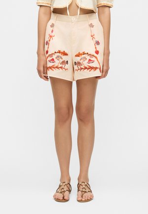 Short beige taille haute avec broderies colorées de fleurs et de champignons, porté avec des sandales marron à brides.