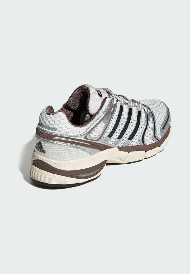 Weißer und metallic silberner Sportschuh mit einem strukturierten Mesh-Obermaterial, das braune Akzente und schwarze Streifenüberzüge an der Seite aufweist.