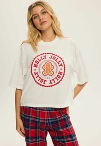 Biała koszulka z krótkim rękawem z czerwoną okrągłą grafiką przedstawiającą logo "HOLLY JOLLY" oraz motywem ciastka piernikowego. Połączona z czerwonymi, niebieskimi i białymi spodniami w kratę.