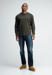 Mann steht mit einem olivgrünen Langarmhemd mit Text, blauen Jeans und braunen Wildlederschuhen vor einem schlichten hellen Hintergrund.