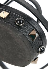 Fond rond d’un sac en cuir noir texturé avec des clous pyramides argentés et des clous noirs en forme de dôme sur fond blanc.
