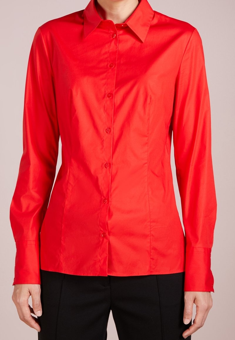 Femme portant une chemise rouge ajustée à manches longues avec col et boutons, associée à un pantalon noir, montrée du cou jusqu'aux hanches.