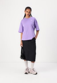 Tricou oversized lavandă, fustă neagră până la genunchi cu buzunare, ciorapi sport albi, teniși roz deschis și o geantă mică neagră.
