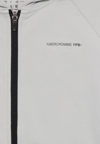 Light gray zip-up hoodie made of soft, stretchy fabric with a black zipper. Features "ABERCROMBIE YPB" logo on the front.