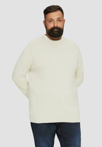 Cremefarbenes Strickpullover mit rundem Halsausschnitt, langen Ärmeln und lockerer Passform, kombiniert mit dunklen Jeans. Schlichtes, strukturiertes Design.