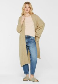 Hellbrauner, übergroßer Cardigan aus weichem Stoff, getragen über einem hellen Oberteil und blauen Jeans. Hat eine offene Vorderseite und weite Ärmel.