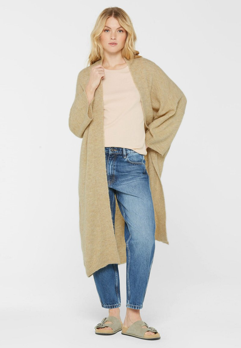 Hellbrauner, übergroßer Cardigan aus weichem Stoff, getragen über einem hellen Oberteil und blauen Jeans. Hat eine offene Vorderseite und weite Ärmel.