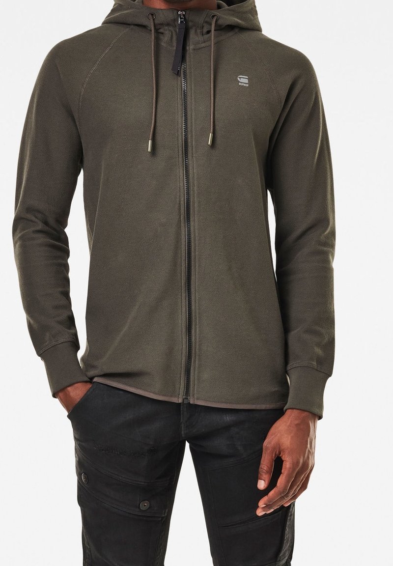 Homme portant un sweat à capuche zippé vert olive avec cordons et un pantalon cargo noir, debout, main gauche dans la poche et main droite détendue.