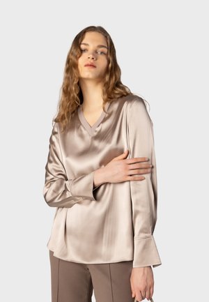 Kvinde med bølget hår iført en lysebeige satinbluse med lange ærmer og brune bukser, poserer mod en ensfarvet lys baggrund.