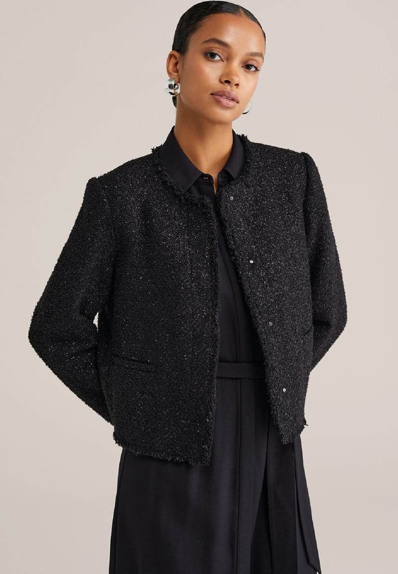 Naine, kellel on rõngas kõrvarõngad, kannab tekstuurset musta lühikest jakki musta kraedressi peal, seistes käed selja taga.