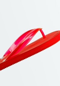 Tongs en caoutchouc rouge avec des brides rouges translucides et brillantes, vue latérale en angle sur un fond clair uni.