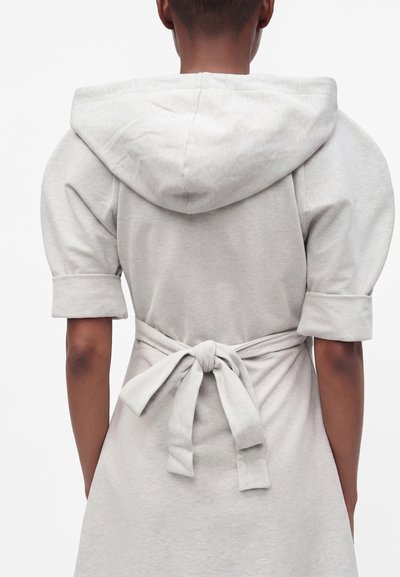 Personne portant un vêtement à capuche gris clair, manches retroussées et ceinture nouée à la taille, vue de dos sur un fond uni.