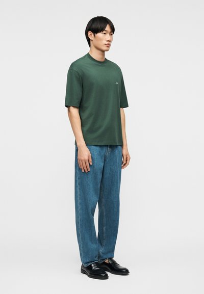 Άνδρας που στέκεται, φοράει κοντομάνικο μπλουζάκι σκούρου πράσινου χρώματος, φαρδιά μπλε τζιν και μαύρα δερμάτινα παπούτσια, μπροστά σε απλό φωτεινό φόντο.