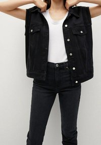 Chaleco de denim negro con cuello, dos bolsillos en el pecho y cierres de botón plateados, llevado sobre una camiseta blanca y combinado con unos vaqueros negros de cintura alta.