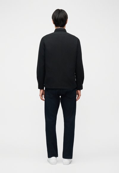 Άτομο που στέκεται γυρισμένο από πίσω, φοράει μαύρο πουκάμισο μακρυμάνικο, σκουρόχρωμα τζιν και λευκά αθλητικά παπούτσια σε απλό ανοιχτό φόντο.