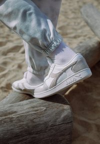 Bílé tenisky s šedými akcenty a texturovaným materiálem, se znakem na boku. Nosené s bledými elastickými joggery se staženými nohavicemi a bílými ponožkami.