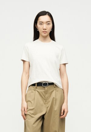 Ungdom med rett svart hår iført en enkel hvit kortermet skjorte, beige plissert bukse og svart belte mot en lys bakgrunn.