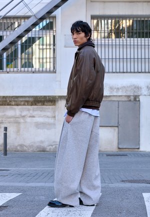 Giacca bomber in pelle marrone con polsini a costine, pantaloni sportivi grigi oversize e scarpe nere, in piedi davanti a uno sfondo urbano.
