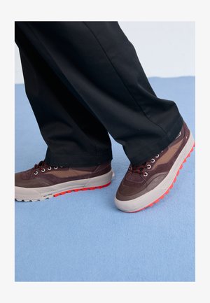 Pruunid ja hallid spordijalatsid tekstuurse pinnaga, punase kummist tallaga ja paeltega. Koos mustade pükstega helesinise tausta ees.