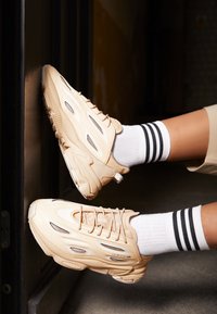 Béžové tenisky Adidas nošené s bílými ponožkami se třemi černými pruhy, nohy ohnuté a chodidla opřená o tmavou svislou plochu.