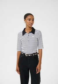 Randig polo t-shirt i vit och svart bomull, med svart krage, korta �ärmar och knäppning fram. Bärs med svarta högmidjade jeans.