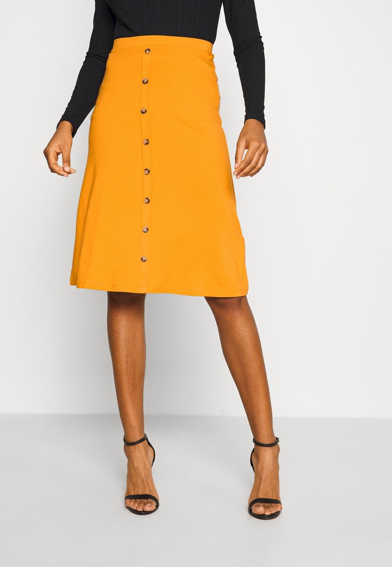 Vrouw draagt een oranje rok tot op de knie met knopen aan de voorkant, gecombineerd met een aansluitende zwarte top met lange mouwen en zwarte pumps met een enkelbandje.