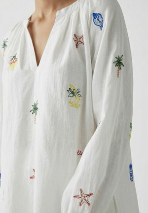 Chemise blanche à manches longues avec des motifs brodés colorés de palmiers, étoiles de mer, coquillages et fruits sur le devant et les manches.