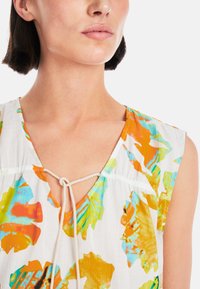 Bluse mit Blumenmuster, die lebendige orangefarbene, gelbe und türkisfarbene Blätter auf einem weißen Hintergrund zeigt, mit einer Schlitz-Krawatten-Detail am Halsausschnitt.