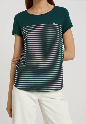 Vrouw die een donkergroen T-shirt met korte mouwen draagt met dunne witte horizontale strepen en een klein wit hartje bij de linkerschouder, gecombineerd met een witte broek.