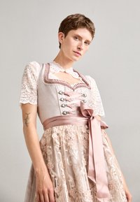 Jasnoróżowy dirndl z koronkowym wykończeniem u góry, srebrnymi dodatkami oraz satynowym paskiem. Spódnica z kwiatowym haftem na przezroczystym materiale.