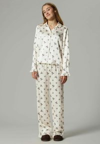 Jonge vrouw in een crème kleurige pyjamastel met kleine zwarte strikprints, staande tegen een effen lichtgrijze achtergrond.