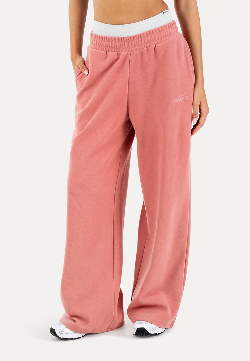 Osoba ubrana w luźne, różowe, wysokoprofilowe spodnie dresowe z bocznymi kieszeniami, w połączeniu z białymi trampkami na białym tle.