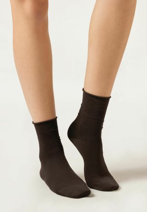 Piernas con calcetines negros hasta el tobillo, un pie plano y el otro levantado sobre los dedos en un fondo claro y simple.