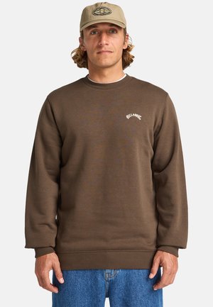 Sudadera de forro polar marrón con puños y dobladillo acanalados, que presenta un logotipo bordado en blanco. Combinada con jeans de mezclilla azul y una gorra beige.